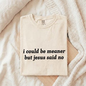 May include: Cream-colored t-shirt with the text "i could be meaner but jesus said no" in black. The shirt is folded on a wooden board, with a neutral-toned blanket in the background. The shirt has a crew neck and short sleeves.