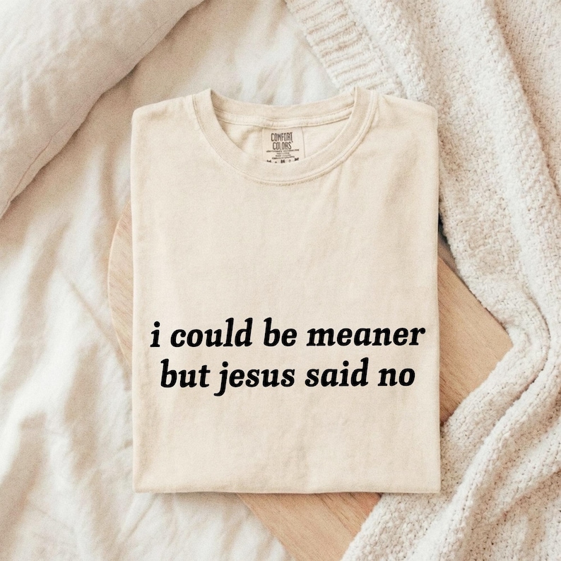May include: Cream-colored t-shirt with the text "i could be meaner but jesus said no" in black. The shirt is folded on a wooden board, with a neutral-toned blanket in the background. The shirt has a crew neck and short sleeves.