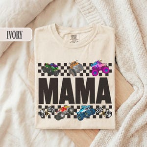 May include: Ivory-colored t-shirt with the word "MAMA" in large, dark letters. Above and below the word are colorful monster truck graphics and checkered flag designs. The shirt is made of a soft material.
