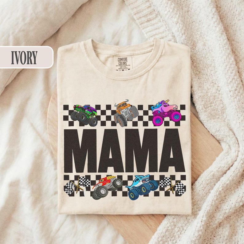 May include: Ivory-colored t-shirt with the word "MAMA" in large, dark letters. Above and below the word are colorful monster truck graphics and checkered flag designs. The shirt is made of a soft material.
