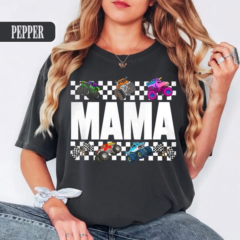 May include: Dark gray t-shirt with the word "MAMA" in large white letters. Above and below the word are checkered flag patterns. The shirt features colorful monster truck graphics in green, orange, pink, red, and blue.
