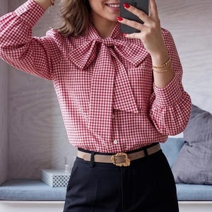 Puede incluir: Una blusa de cuadros rojos y blancos con un gran cuello de lazo. La camisa de manga larga se combina con pantalones negros y un cinturón marrón claro con hebilla dorada. El conjunto es elegante.