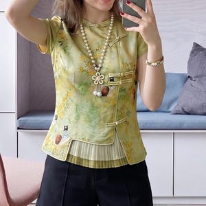 May include: A light green floral print top with a mandarin collar and frog closures. The top is paired with black shorts. A long pearl necklace with a flower pendant and a gold bracelet are also visible.