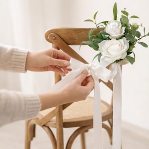 Puede incluir: Una silla de madera adornada con rosas blancas y vegetación, atada con una cinta de satén blanco. La silla tiene un asiento tejido y está siendo decorada con el arreglo floral. El fondo es de un color suave y neutro.