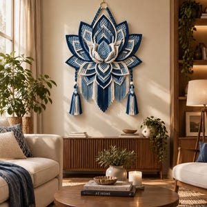 Puede incluir: Un tapiz de macramé grande y complejo con forma de flor de loto, que presenta pétalos en capas en tonos azul marino, azul claro y crema. El diseño incluye detalles de cordón trenzado, dos borlas decorativas largas y una anilla para colgar.