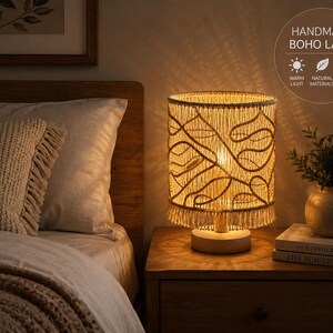 May include: A cylindrical table lamp featuring a beige macrame shade with an intricate knotted pattern and a bottom fringe. The lamp has a circular wooden base and emits a warm glow. Text on the image reads: HANDMADE BOHO LAMP, WARM LIGHT, NATURAL MATERIALS, COZY AMBIANCE.