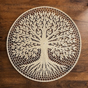 May include: A circular beige crochet doily featuring an intricate tree of life design. The pattern displays a central trunk that branches out into detailed leaves and roots, all connected by a fine, openwork mesh structure made of thin yarn.