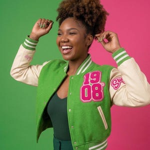Op de afbeelding: Groene en crèmekleurige varsity jas met het nummer "1908" in roze op de voorkant. De jas heeft crèmekleurige mouwen en groen-wit gestreepte manchetten. De jas wordt over een groene top gedragen.