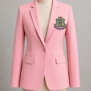 May include: A light pink blazer with a single-button closure and two front pockets. The blazer features a colorful embroidered crest on the left chest. The crest includes the letters AKA and other text.