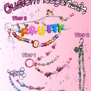 May include: Pink background with the text "Custom Keychain". Several beaded keychains are displayed, including one spelling "FRUITY" with colorful letter beads. Other keychains feature names and phrases like "PROTECT THE DOLLS".