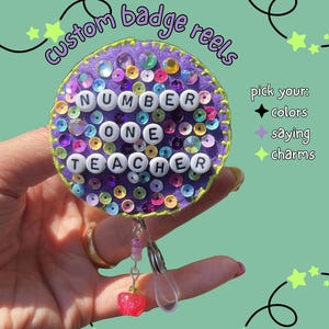 May include: A custom badge reel with the text "NUMBER ONE TEACHER" in white beads. The purple felt reel is decorated with colourful sequins and a small charm. The image also includes the text "custom badge reels" and "pick your: colours, saying, charms."