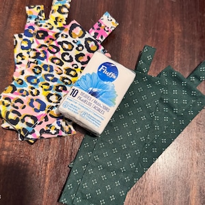 Peut inclure: Un paquet de mouchoirs Fluffs avec un motif de fleur bleue, placé à côté de plusieurs bandes de tissu. Un ensemble de bandes présente un imprimé léopard multicolore avec du rose, du bleu et du jaune, tandis que l'autre est vert foncé avec un petit motif blanc.