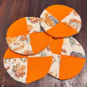 Könnte beinhalten: Drei runde Stoffuntersetzer mit einem Patchwork-Design aus abwechselnden einfarbigen orangefarbenen Segmenten und cremefarbenem Stoff, bedruckt mit herbstlichen Motiven wie Ahornblättern, Donuts, Kaffeetassen und dem Wort pumpkin.