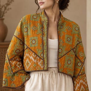 May include: A cropped, open-front jacket featuring a quilted texture with a mustard yellow and green geometric pattern. The garment has wide sleeves, a mandarin-style collar, and dark brown piping along the edges and seams, worn over a white top.