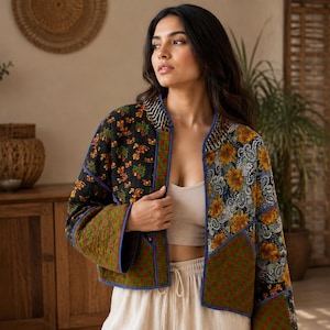 May include: A quilted jacket featuring a patchwork design with floral and paisley patterns in black, yellow, green, and blue. The jacket has long sleeves, a collarless neckline, and blue trim along the edges, worn over a beige crop top and pants.