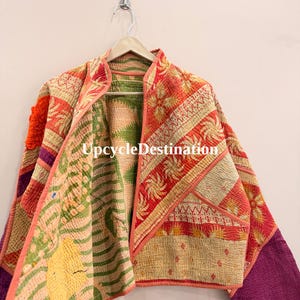 May include: A quilted jacket featuring a patchwork design with orange, green, yellow, and purple fabric panels. The garment has a mandarin collar, long sleeves, and visible kantha stitching throughout. The text UpcycleDestination is visible.