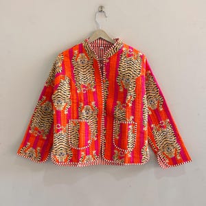 May include: A reversible quilted jacket with a vibrant tiger print. The jacket features a bold orange and pink striped pattern with tiger motifs. It has a mandarin collar, two pockets, and a red and white striped lining. The jacket is hanging on a wooden hanger.