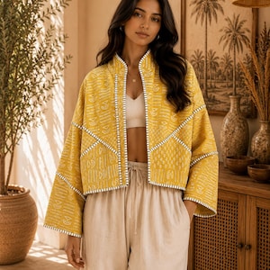 Peut inclure: Une veste courte jaune moutarde avec un motif géométrique blanc, un col montant et des manches larges. La veste est agrémentée de surpiqûres blanches le long des coutures et des bords, portée sur un haut court côtelé blanc et un pantalon beige.