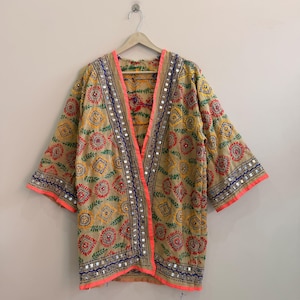 Può includere: Una giacca beige aperta sul davanti con ricami floreali e geometrici multicolori. Il capo è impreziosito da piccoli specchietti riflettenti e da un bordo rosa neon brillante lungo il colletto e le estremità delle maniche.
