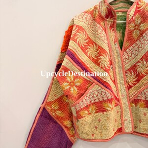 May include: A quilted jacket featuring a patchwork of orange, cream, and purple fabric with floral and geometric patterns. The garment has a mandarin collar, long sleeves, and visible stitching throughout. The text UpcycleDestination is visible.