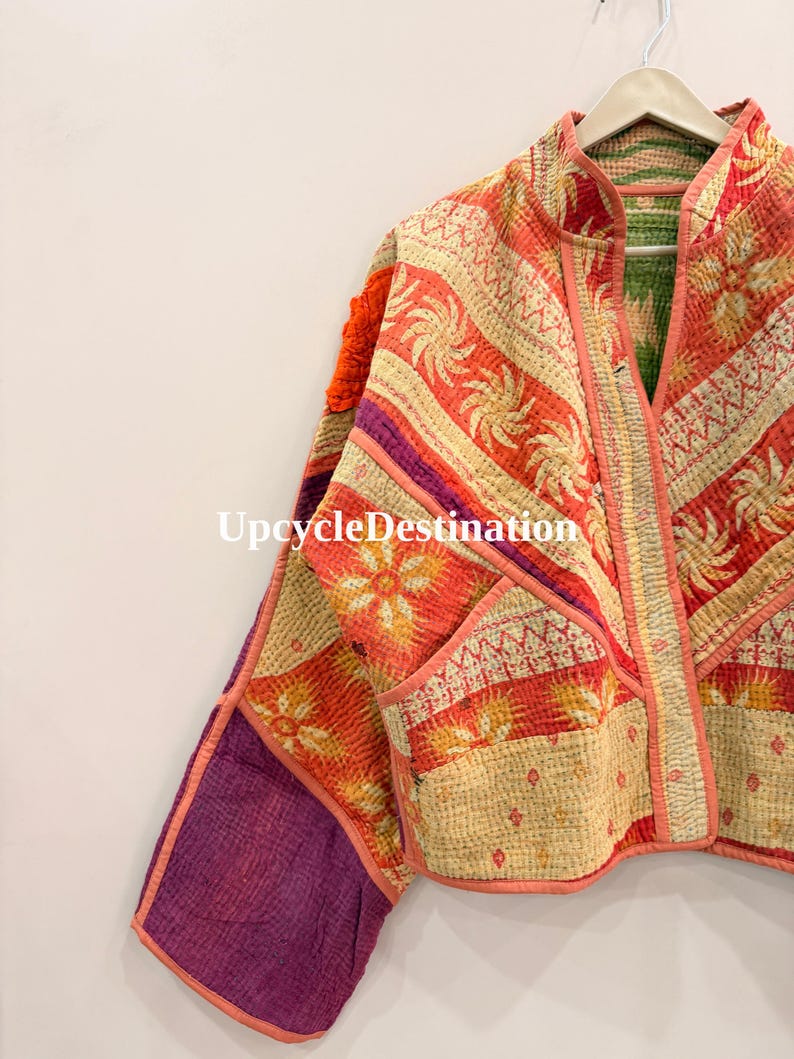 May include: A quilted jacket featuring a patchwork of orange, cream, and purple fabric with floral and geometric patterns. The garment has a mandarin collar, long sleeves, and visible stitching throughout. The text UpcycleDestination is visible.