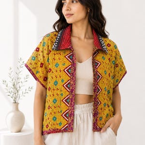 May include: A short-sleeved, open-front jacket in mustard yellow with a quilted texture. It features a vibrant floral pattern in red, blue, and white, with a contrasting zigzag border along the placket and a dark patterned collar.