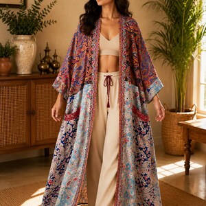 Puede incluir: Un kimono largo abierto por delante con un vibrante estampado de cachemira multicolor en tonos azules, rojos y naranjas. La prenda tiene mangas anchas, dos bolsillos delanteros y un ribete decorativo en contraste a lo largo del cuello.