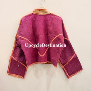 May include: A cropped jacket in a vibrant magenta hue featuring visible quilted stitching throughout. The garment has long sleeves, a high collar, and orange piping along the seams. The text UpcycleDestination is printed in white across the back.