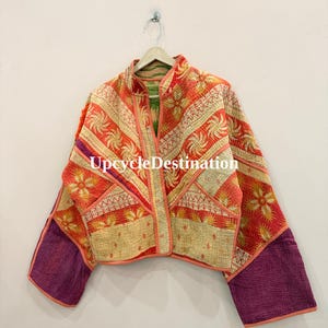 May include: A quilted jacket featuring a vibrant patchwork of orange, red, and cream floral patterns with contrasting solid purple sleeves. The garment has a mandarin collar, front pockets, and the text UpcycleDestination printed in white.