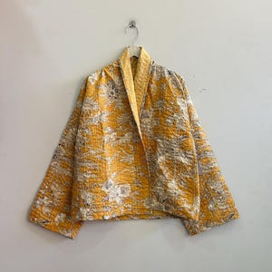 Puede incluir: Una chaqueta acolchada de color amarillo mostaza con un estampado floral blanco en toda la prenda. La chaqueta tiene un cuello ancho, mangas largas y un dobladillo recto. La tela muestra detalles de costuras visibles en toda la superficie.