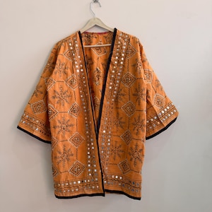 Könnte beinhalten: Eine orangefarbene Jacke mit offener Vorderseite, die mit schwarzen, gestickten geometrischen Mustern und sternartigen Motiven versehen ist. Das Kleidungsstück ist mit kleinen reflektierenden Spiegeln verziert und hat einen schwarzen Saum.