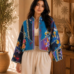 May include: A cropped jacket featuring a patchwork design with vibrant blue, purple, and floral patterns. The garment has long, wide sleeves, a straight front opening, and visible stitching throughout the fabric, paired with beige drawstring trousers.