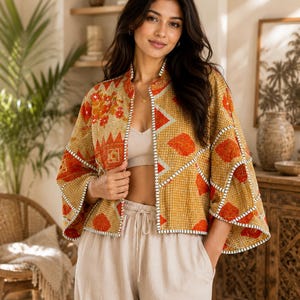 May include: A mustard yellow quilted jacket featuring a vibrant orange and white geometric and floral pattern. The jacket has wide three-quarter length sleeves, a stand-up collar, and visible white stitched detailing along the edges and seams.