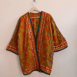 Könnte beinhalten: Eine senfgelbe Jacke mit offener Vorderseite und aufwendigen Stickereien in roten, grünen und blauen kreisförmigen Mustern. Das Kleidungsstück ist mit kleinen reflektierenden Spiegelakzenten verziert und hat einen dunkelgrünen Saum.