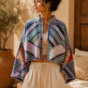 May include: A cropped, open-front jacket featuring a patchwork design of quilted fabric in various shades of blue, purple, and green with diagonal striped patterns. The garment has wide, three-quarter length sleeves and a light-colored trim along the edges.