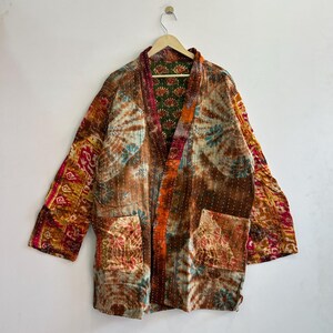 Könnte beinhalten: Eine langärmelige Jacke im Kimono-Stil aus Patchwork-Stoff mit einer Mischung aus braunen, orangen und blaugrünen Batikmustern. Das Kleidungsstück verfügt über zwei Vordertaschen, ein dunkelgrünes Innenfutter mit Paisley-Muster und weiße Steppnähte.