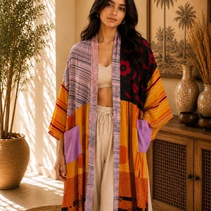 May include: A long, open-front kimono featuring a patchwork design with various patterns and colors, including mustard yellow, black, lavender, and grey stripes. The garment has two large lavender patch pockets and a contrasting patterned trim.