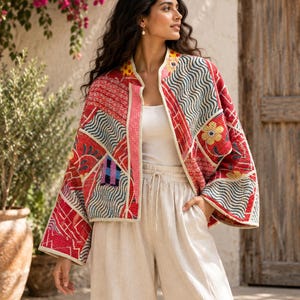 May include: A quilted jacket featuring a patchwork design with red, white, and blue patterns, including wavy lines and floral motifs. The jacket has an open front, wide sleeves, and a cream-coloured trim along the edges, worn over a white top.