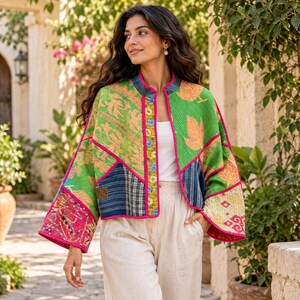 May include: A cropped, open-front jacket featuring a vibrant patchwork design with green, orange, and blue fabric panels. The garment is accented with bright pink trim along the edges and a patterned placket, worn over a plain white top and light trousers.
