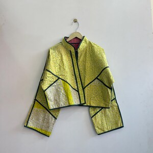 May include: A yellow jacket with a geometric design and black trim hangs on a wooden hanger. The jacket has a high collar and long sleeves. The interior lining is pink.