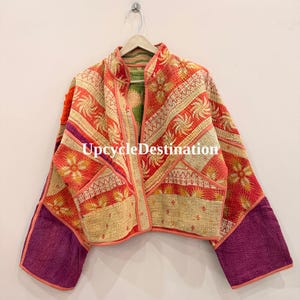 May include: A cropped jacket featuring a patchwork design with orange, red, and cream floral patterns, accented by solid purple sleeves. The garment has a mandarin collar, open front, and visible kantha-style stitching. The text UpcycleDestination is visible.