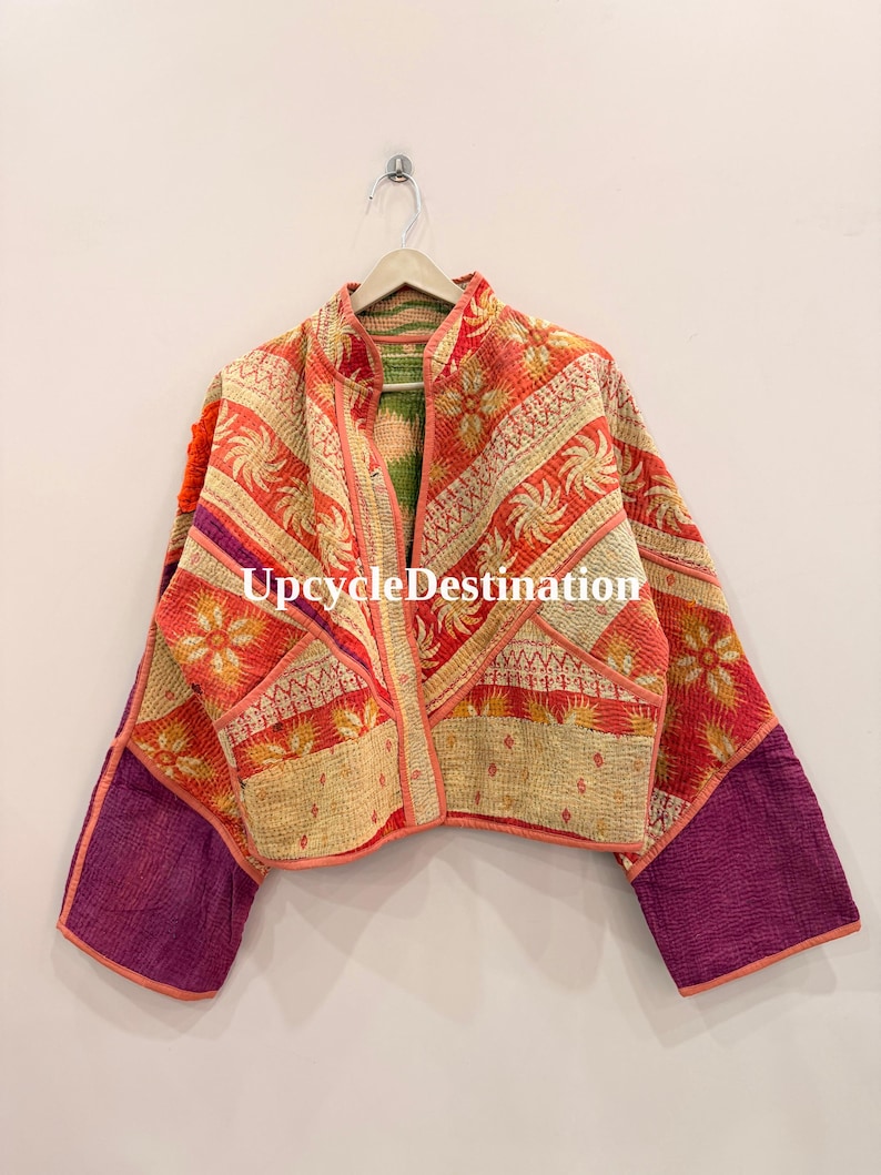 May include: A cropped jacket featuring a patchwork design with orange, red, and cream floral patterns, accented by solid purple sleeves. The garment has a mandarin collar, open front, and visible kantha-style stitching. The text UpcycleDestination is visible.