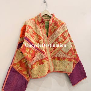 May include: A quilted jacket featuring a patchwork design with orange, cream, and purple fabric panels. The garment displays intricate floral and geometric patterns, a stand-up collar, long sleeves, and the text UpcycleDestination printed centrally.
