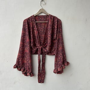 May include: A burgundy paisley print wrap top with long, flared sleeves and ruffled cuffs. The top is tied at the waist with long fabric ties. The garment is hanging on a wooden hanger against a white wall.