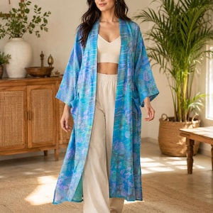 May include: A long, open-front kimono robe featuring a vibrant blue and teal watercolor-inspired floral pattern. The garment has wide, three-quarter length sleeves, a relaxed silhouette, and two front patch pockets, worn over a cream-colored crop top.