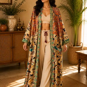 Op de afbeelding: Een lange, aan de voorzijde open kimono met een levendig, meerkleurig paisley-patroon in tinten oranje, goud, zwart en blauwgroen. Het kledingstuk heeft wijde mouwen, twee opgestikte zakken aan de voorzijde en een decoratieve rand langs de opening.