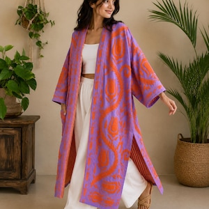 Può includere: Un lungo kimono aperto sul davanti di colore viola chiaro con un vivace motivo floreale arancione. Il capo presenta maniche larghe, spacchi laterali e un orlo dritto. È indossato sopra un top corto bianco e pantaloni bianchi a gamba larga.
