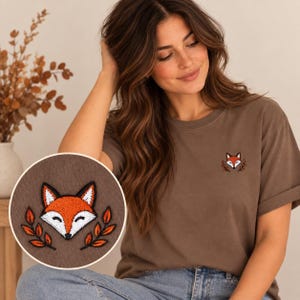 Könnte beinhalten: Ein braunes kurzärmeliges T-Shirt mit einem kleinen gestickten Aufnäher auf der linken Brust. Der Aufnäher zeigt einen stilisierten Fuchskopf in Orange, Weiß und Schwarz, flankiert von zwei kleinen orangefarbenen Blattzweigen auf beiden Seiten.
