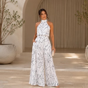 May include: White halter-neck jumpsuit with a black abstract design. The wide-leg jumpsuit features a high neckline, pockets, and a matching print. A small clutch is held in the hand. The outfit is suitable for a special occasion.