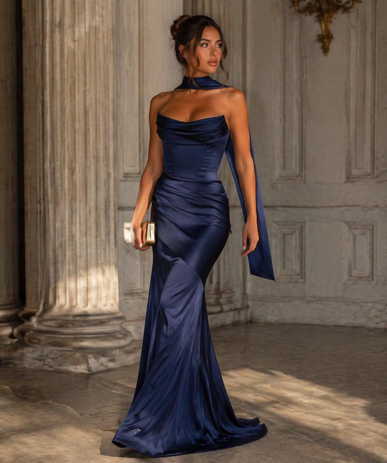 May include: A navy blue, floor-length gown with a draped bodice and a flowing skirt. The dress features a halter neckline and a long, flowing panel. A small, gold clutch is held in the hand.